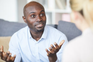 Puzzled young black man with bald head talking to psychologist and gesturing hands while expressing his emotions at therapy session