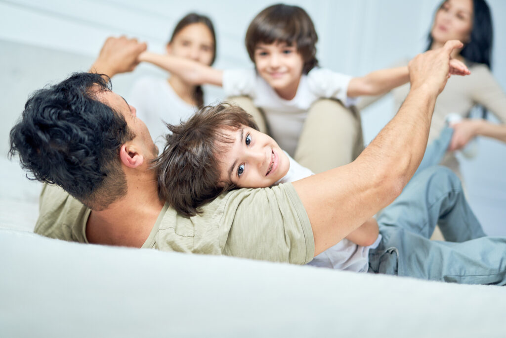 Portrait of cheerful little latin boy smiling at camera, playing with his parents and siblings on a bed at home. Happy childhood, parenthood concept